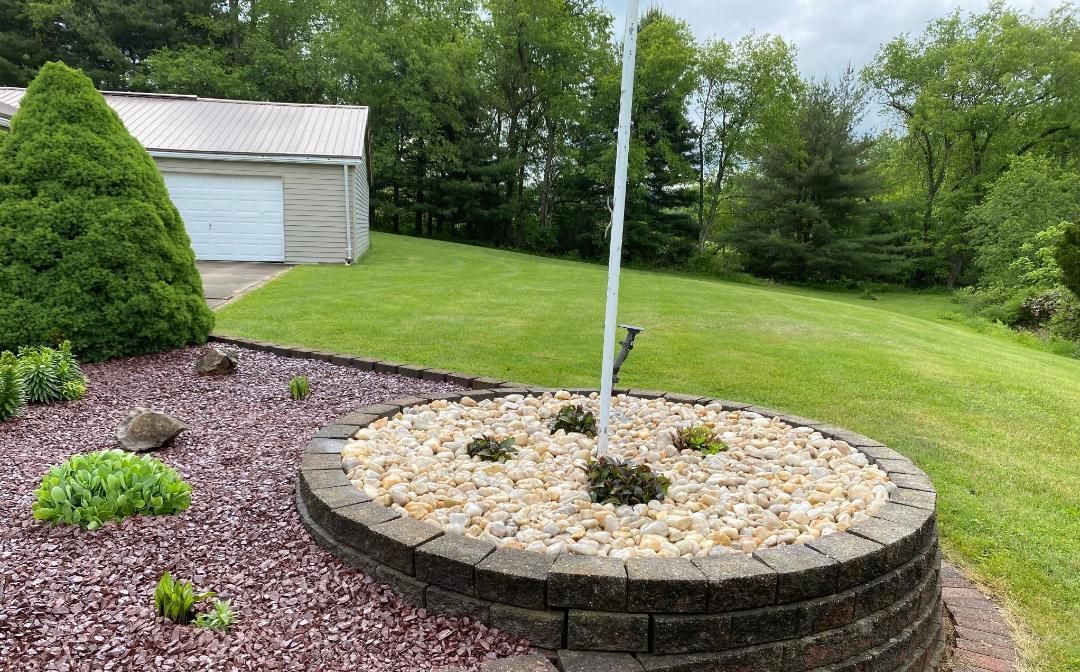 Oval-shaped garden bed with tan rocks, small plants, and a flagpole; grassy lawn and garage in background.