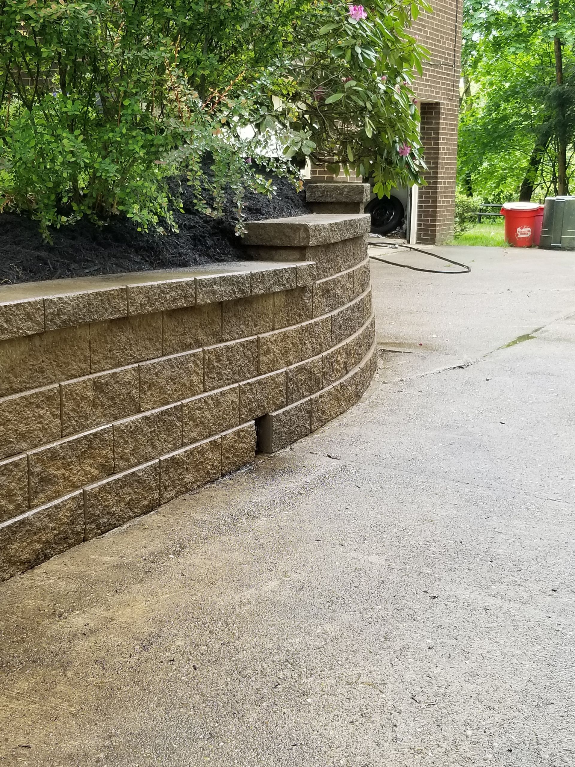 Curved retaining wall made of brown blocks bordering a driveway, with mulch and greenery above.
