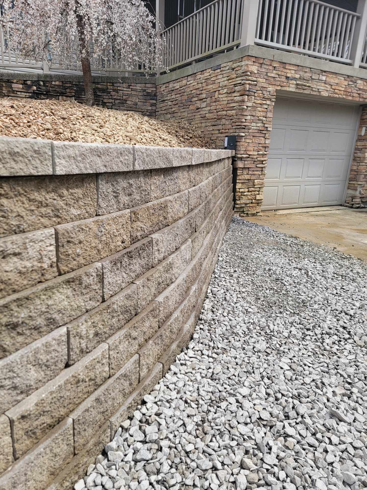 A retaining wall made of beige blocks curves next to a gravel path, a garage and part of a house.
