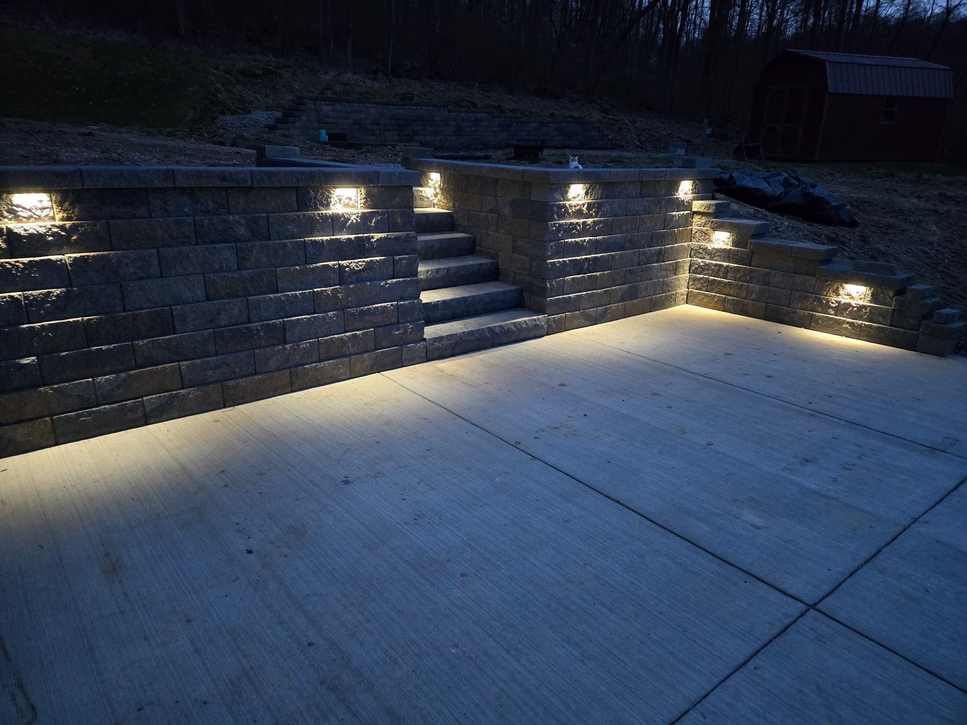 Nighttime outdoor concrete patio with lit stone retaining walls and stairs.