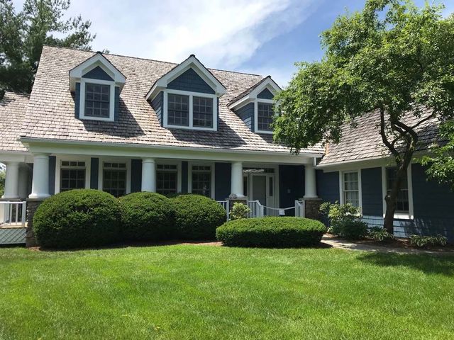 Green suburban house with dormer windows, white porch columns, trimmed hedges, and a front lawn