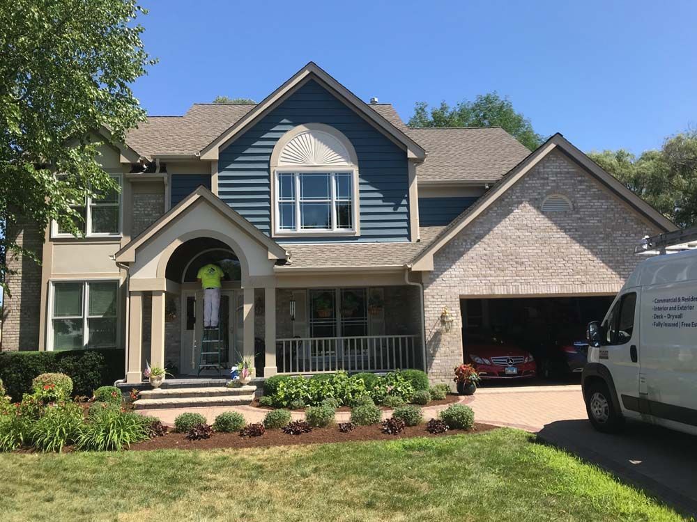 Suburban two-story house with gray siding, brick facade, front porch, and open garage on a sunny day