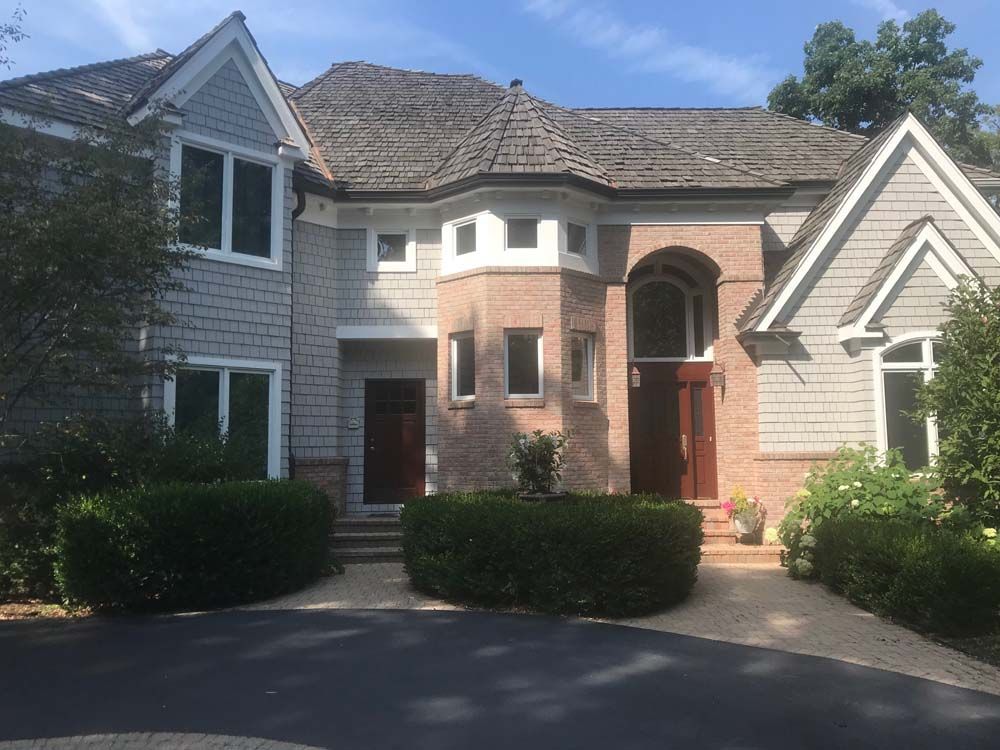 Large two-story brick and siding house with arched entryway, driveway, and trimmed bushes under sunny skies