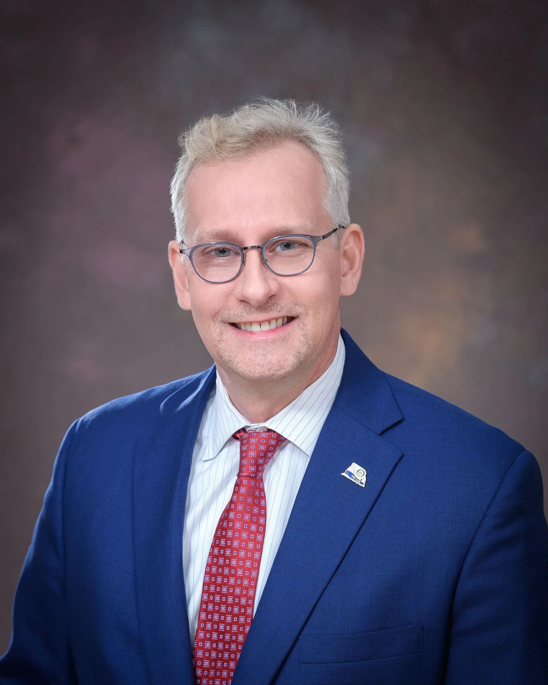 Headshot of a smiling person in a blue suit, red tie, and glasses against a studio backdrop