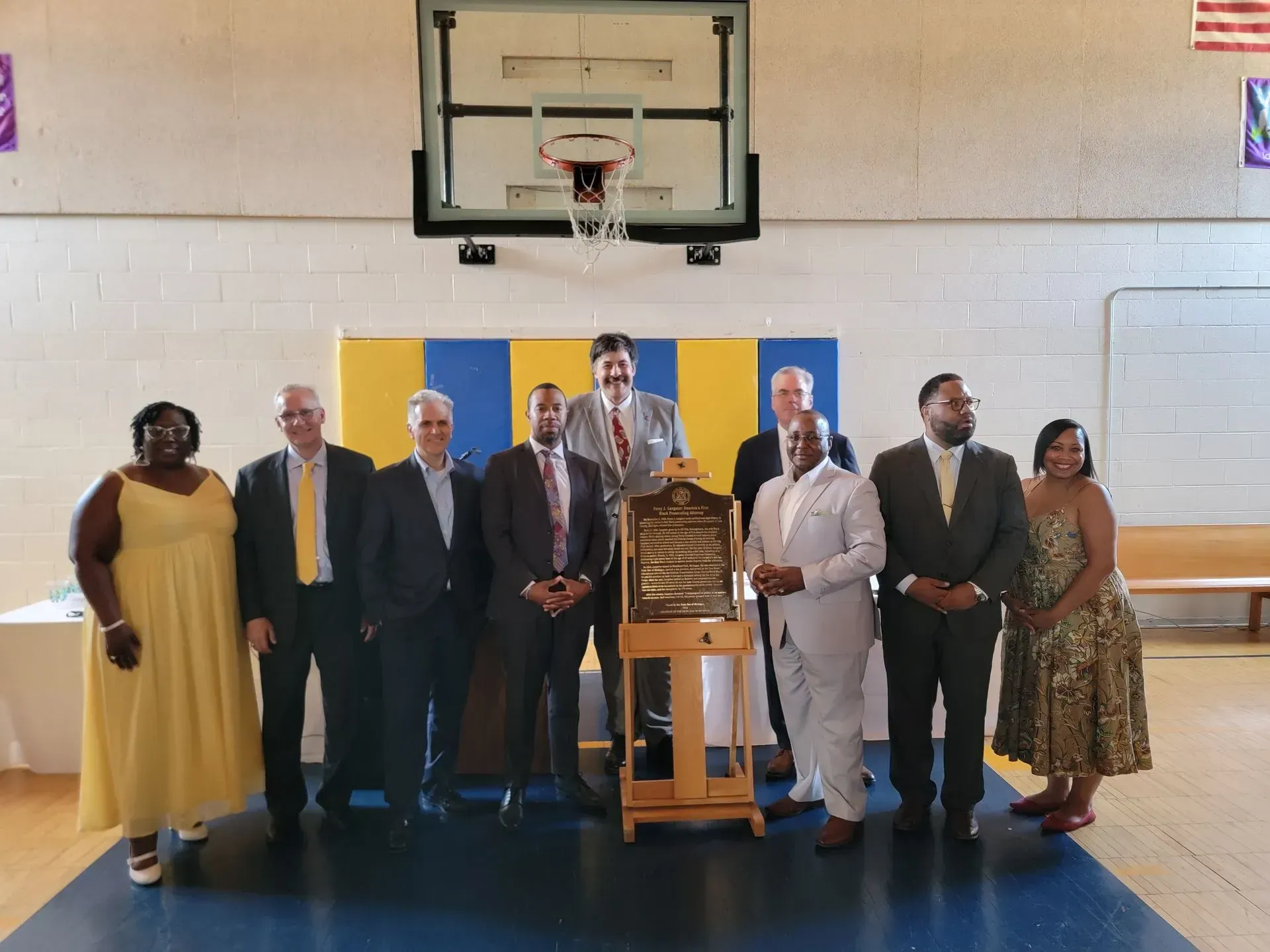 Group of people posing beside an award plaque in a gymnasium under a basketball hoop.