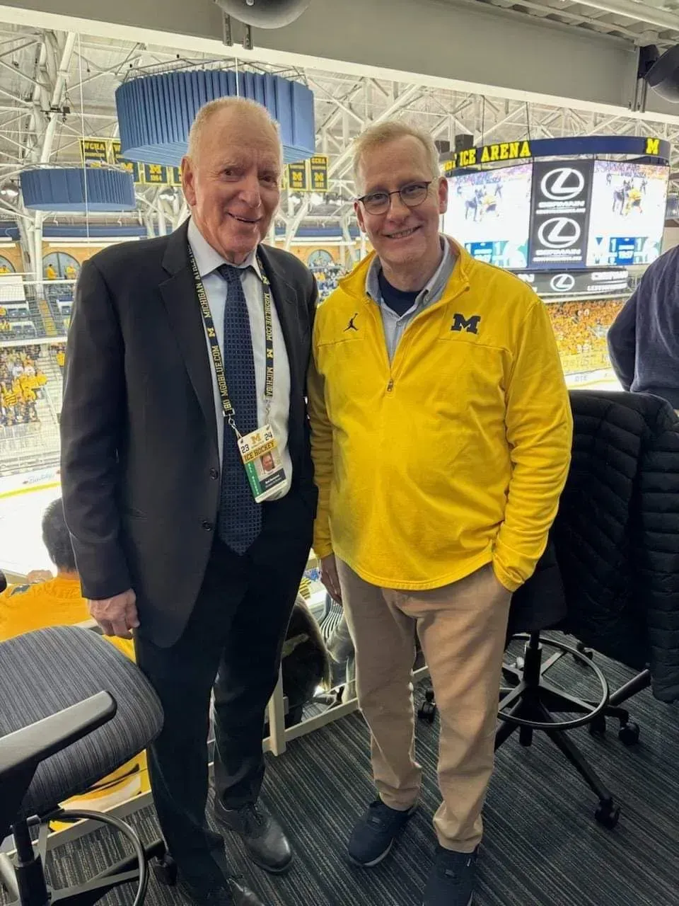 Two men standing in a sports broadcast booth, one in a suit and one in a yellow jacket, smiling.
