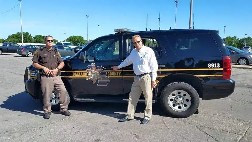 Two men pose beside a black sheriff SUV in a sunny parking lot.
