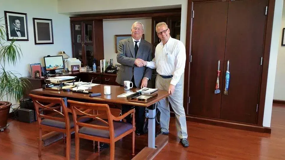 Two men shaking hands in an office with a desk, chairs, wood floors, and dark cabinets.