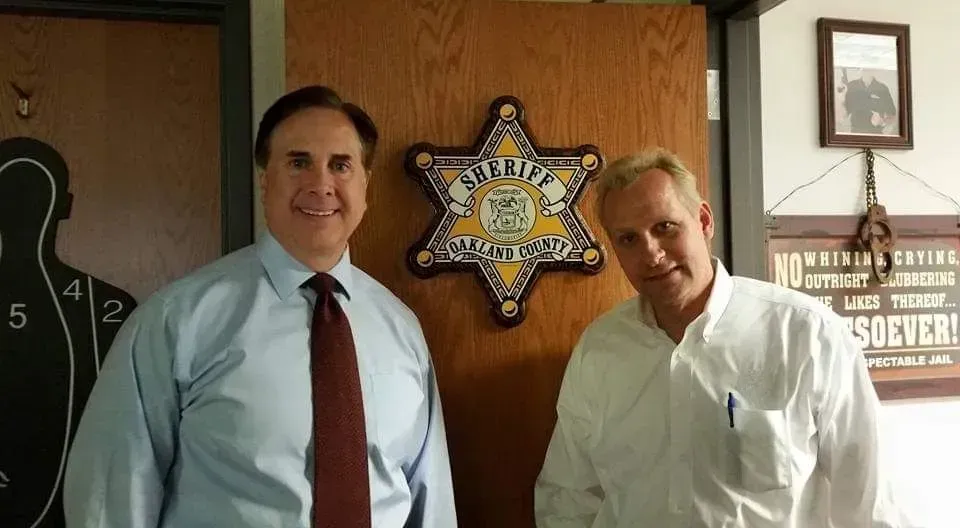 Two men standing in an office beside a sheriff's star plaque and wall sign