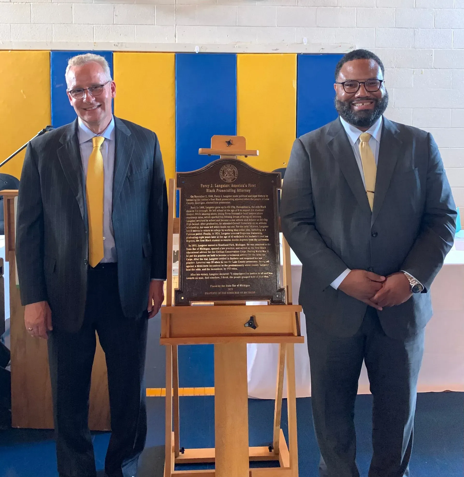Two men in suits stand beside a plaque on a stand in a blue-and-yellow room.