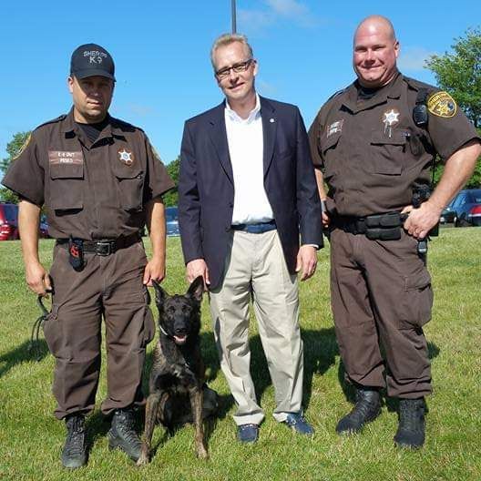 Two sheriff deputies standing with a police dog beside a man in a blazer outdoors on grass.