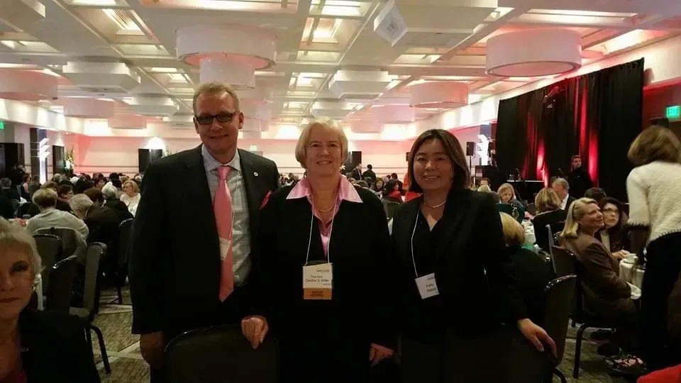 Three people posing at a conference reception in a crowded banquet hall, wearing lanyards.