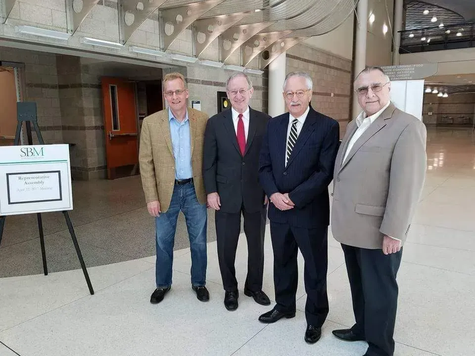 Four men in suits pose indoors beside a sign in a modern lobby.