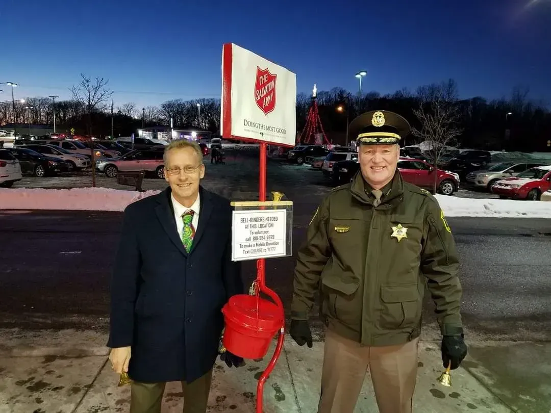 Two men stand beside a red Salvation Army donation sign at dusk in a parking lot.