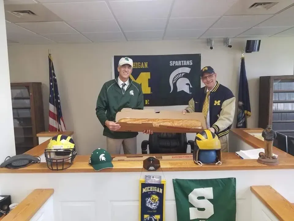 Two men in green and beige jackets holding a wooden plaque at a Michigan State-themed press conference table.
