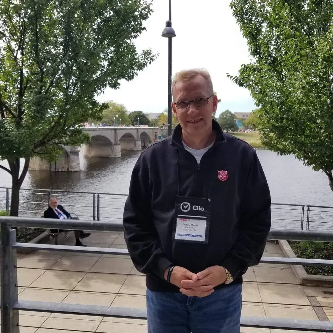 Person standing by a river with a stone bridge in the background, wearing a dark jacket and lanyard.