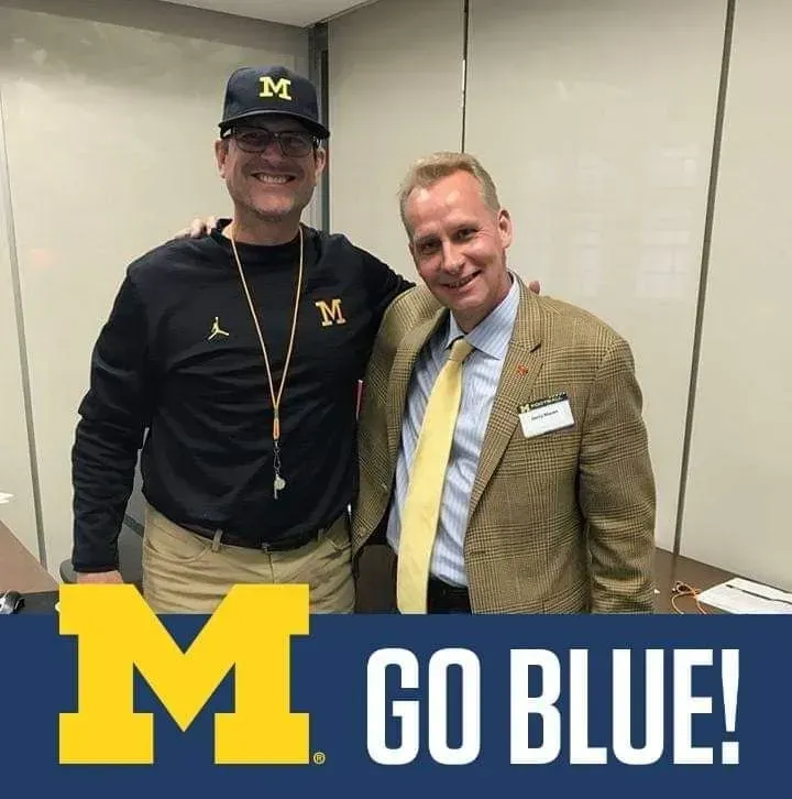 Two men standing indoors, smiling, with a Michigan “M” and “GO BLUE!” text overlay