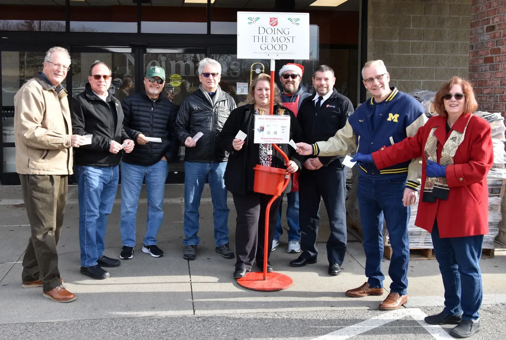 Group of people gathered outdoors around a red stand in front of a building, posing for a photo