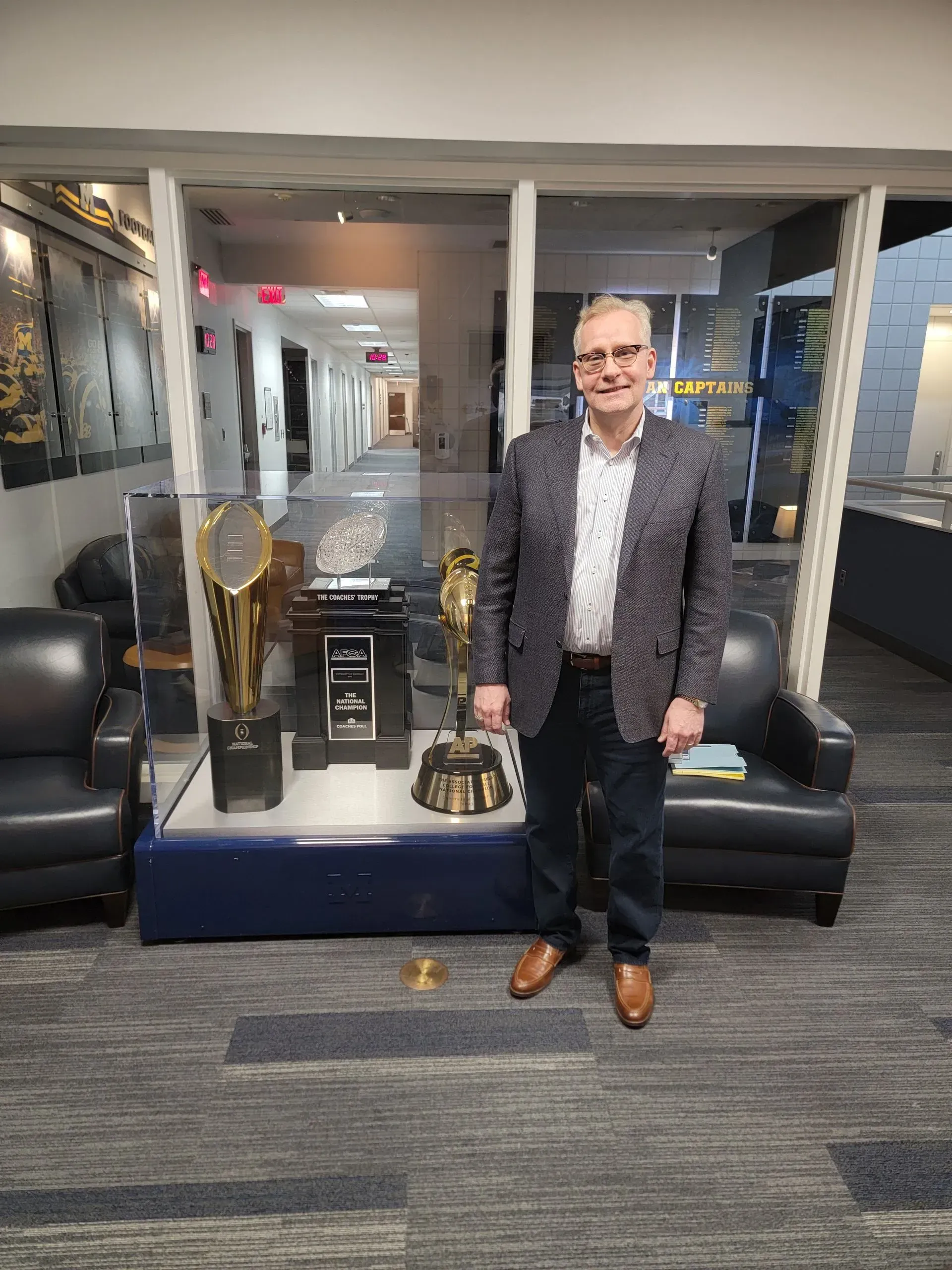 Man standing in a lobby beside display cases with trophies and a large gold cup