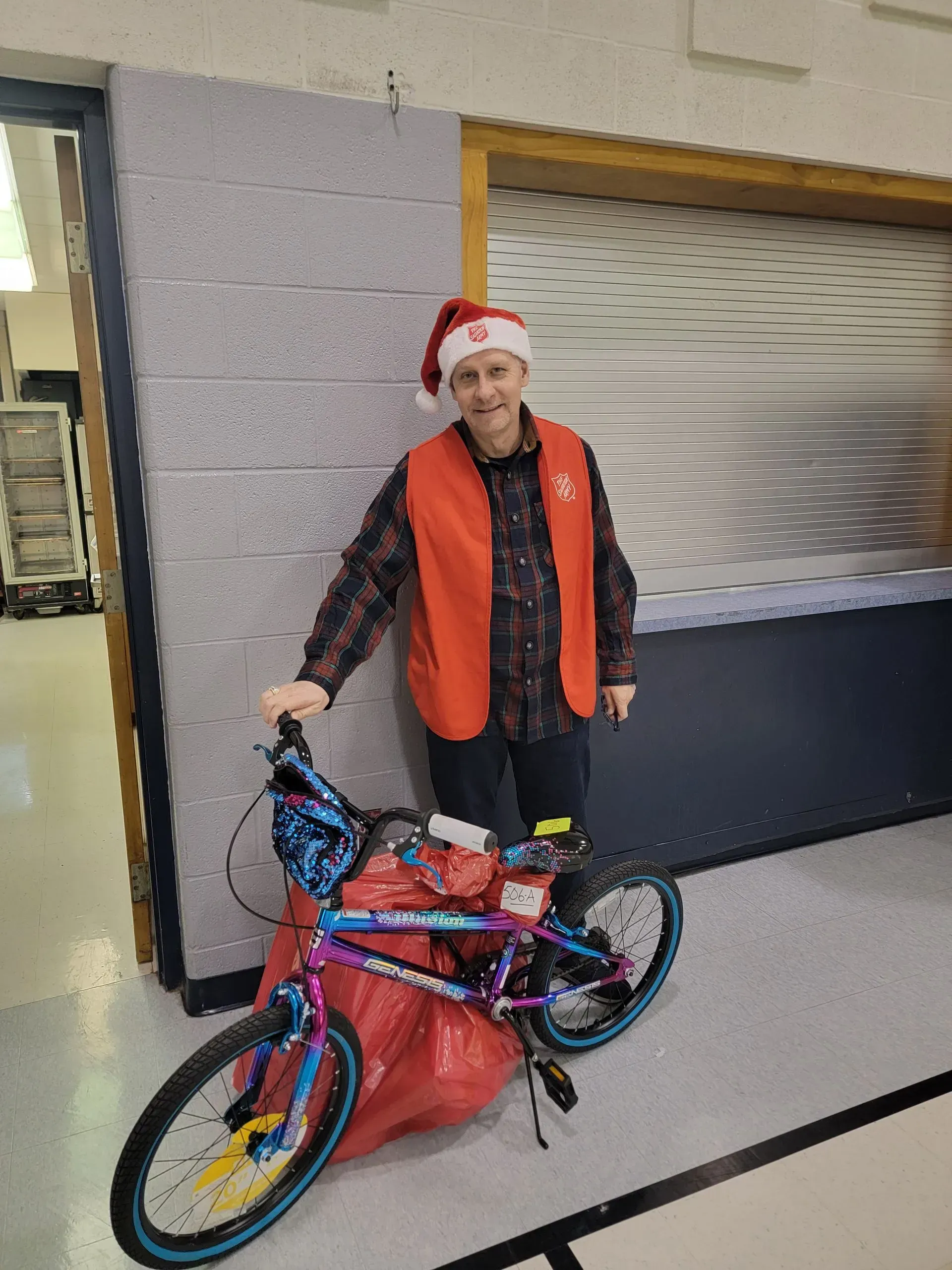 Person in a Santa hat standing indoors beside a decorated bicycle and red bag