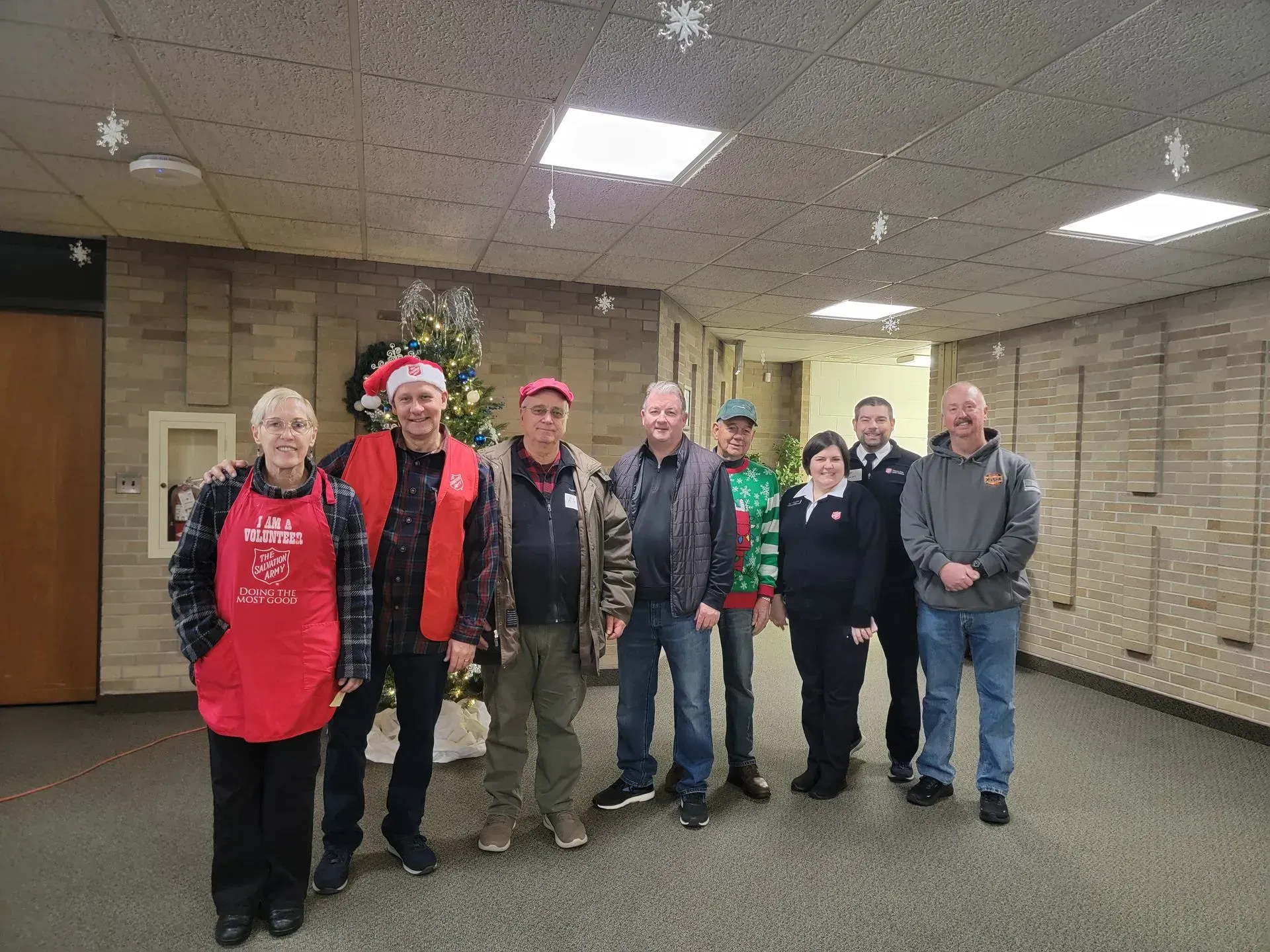 Group of people standing together indoors in a room with beige brick walls, wearing red and dark jackets.