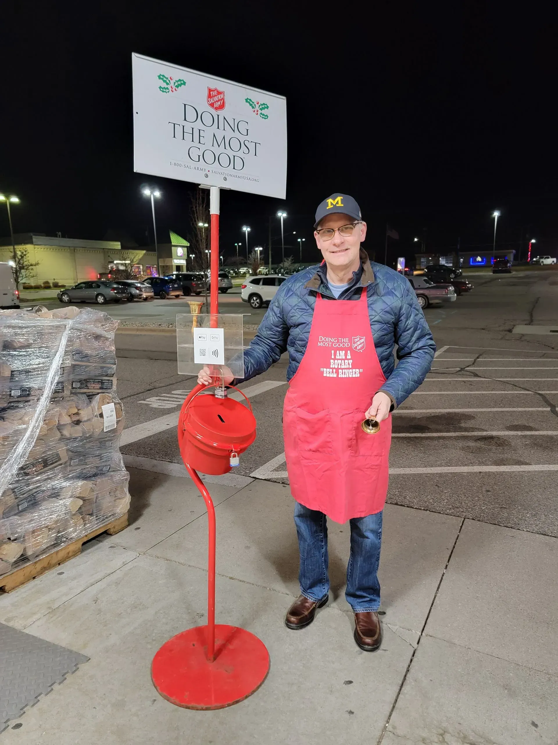Person in red apron stands beside a red donation can in a parking lot at night.