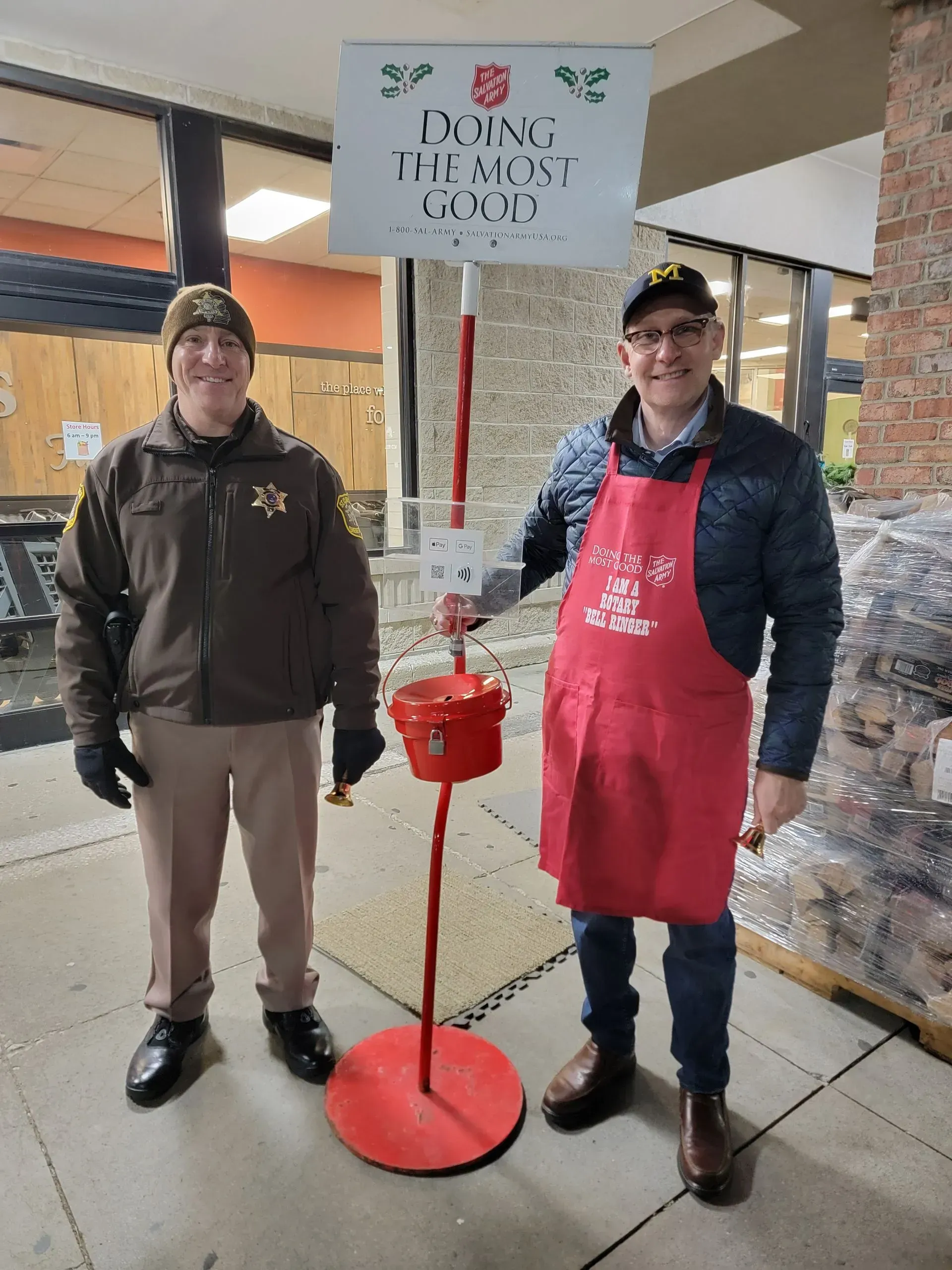 Two people stand beside a red donation bucket under a sign at a covered outdoor shelter.