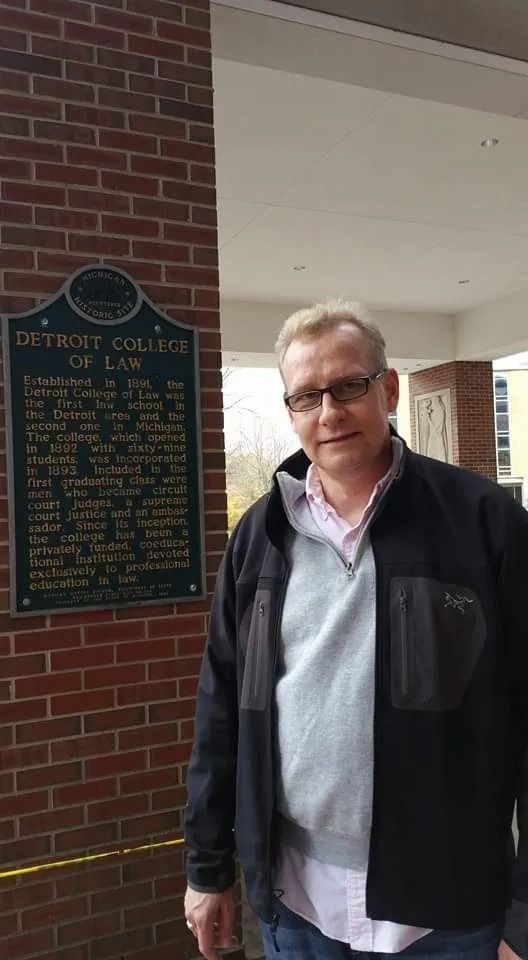 Man standing under a covered brick walkway beside a signboard, wearing a black jacket and light pink shirt.