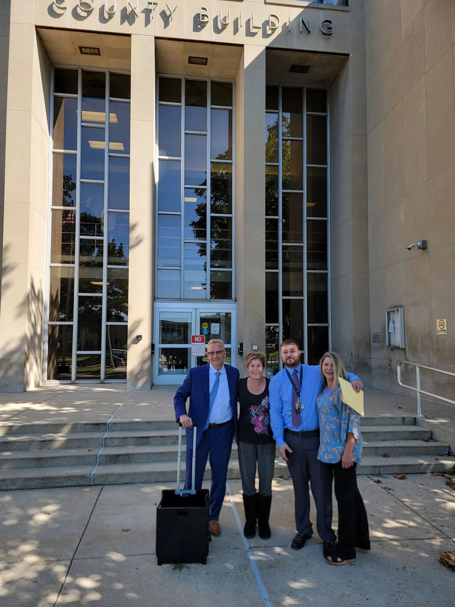 Four people standing outside a county building entrance with tall glass doors and steps.