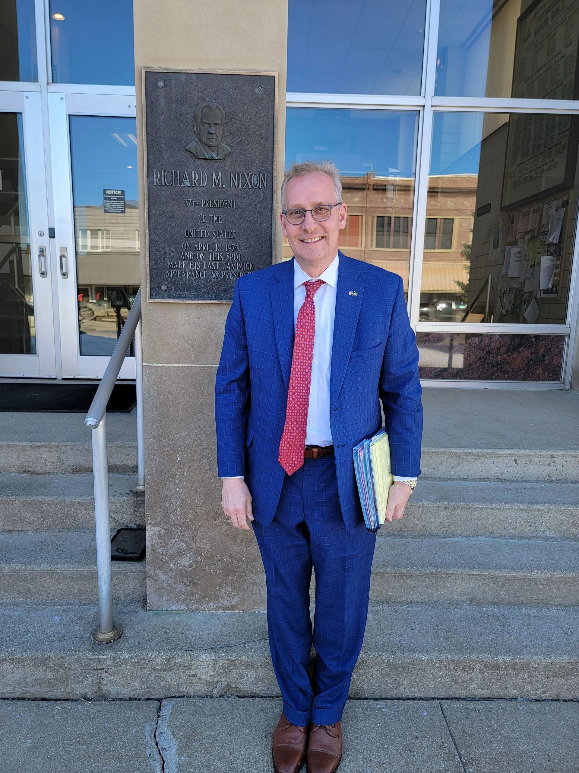 Man in blue suit and red tie standing outside a building entrance, holding a book.
