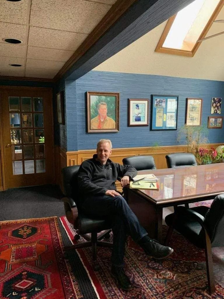 Person seated at a conference table in a wood-paneled office with framed art and skylight.