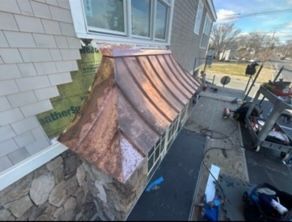 Copper-clad bay window roof under repair beside a stone house wall, with tools and materials on a driveway.