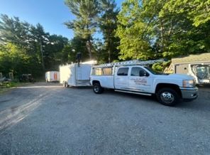 White utility truck with trailer parked on a gravel road surrounded by trees and greenery.