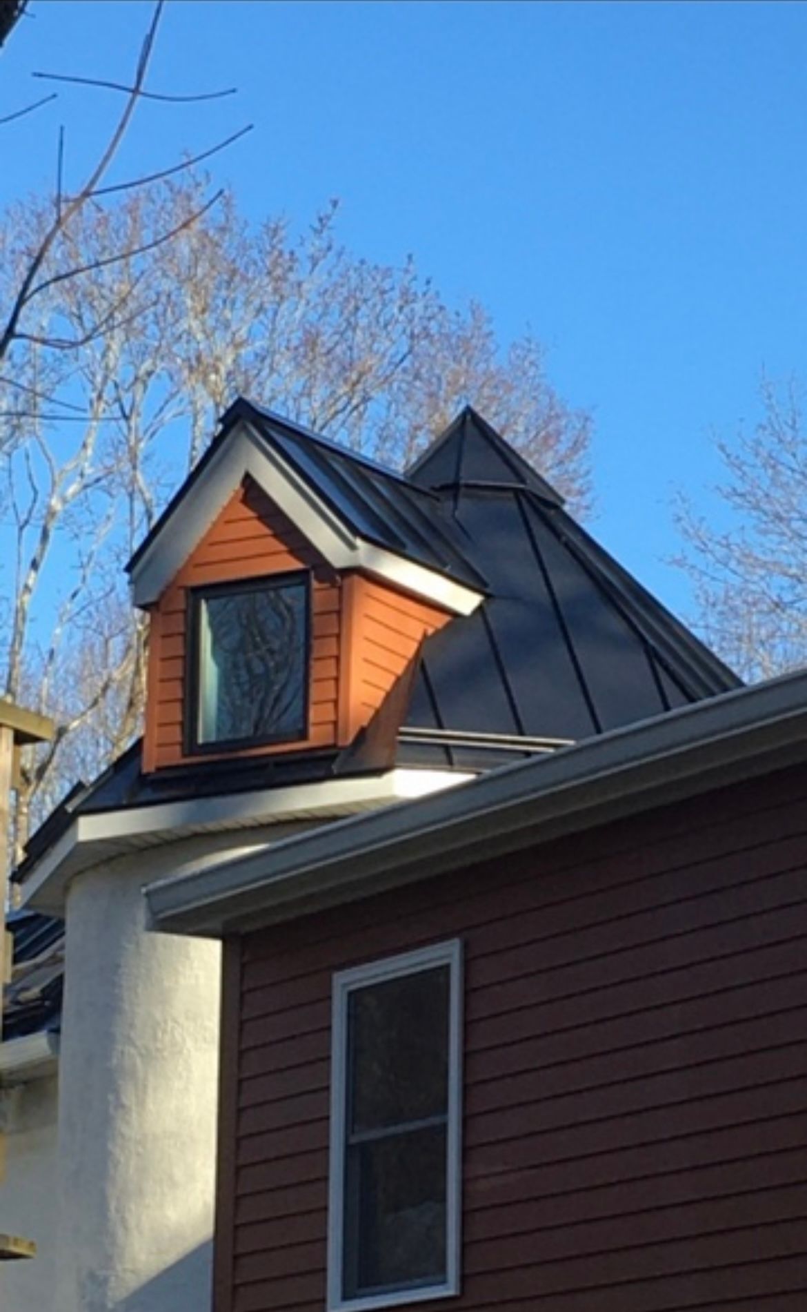 House roof with black metal panels and a small orange dormer against a blue sky