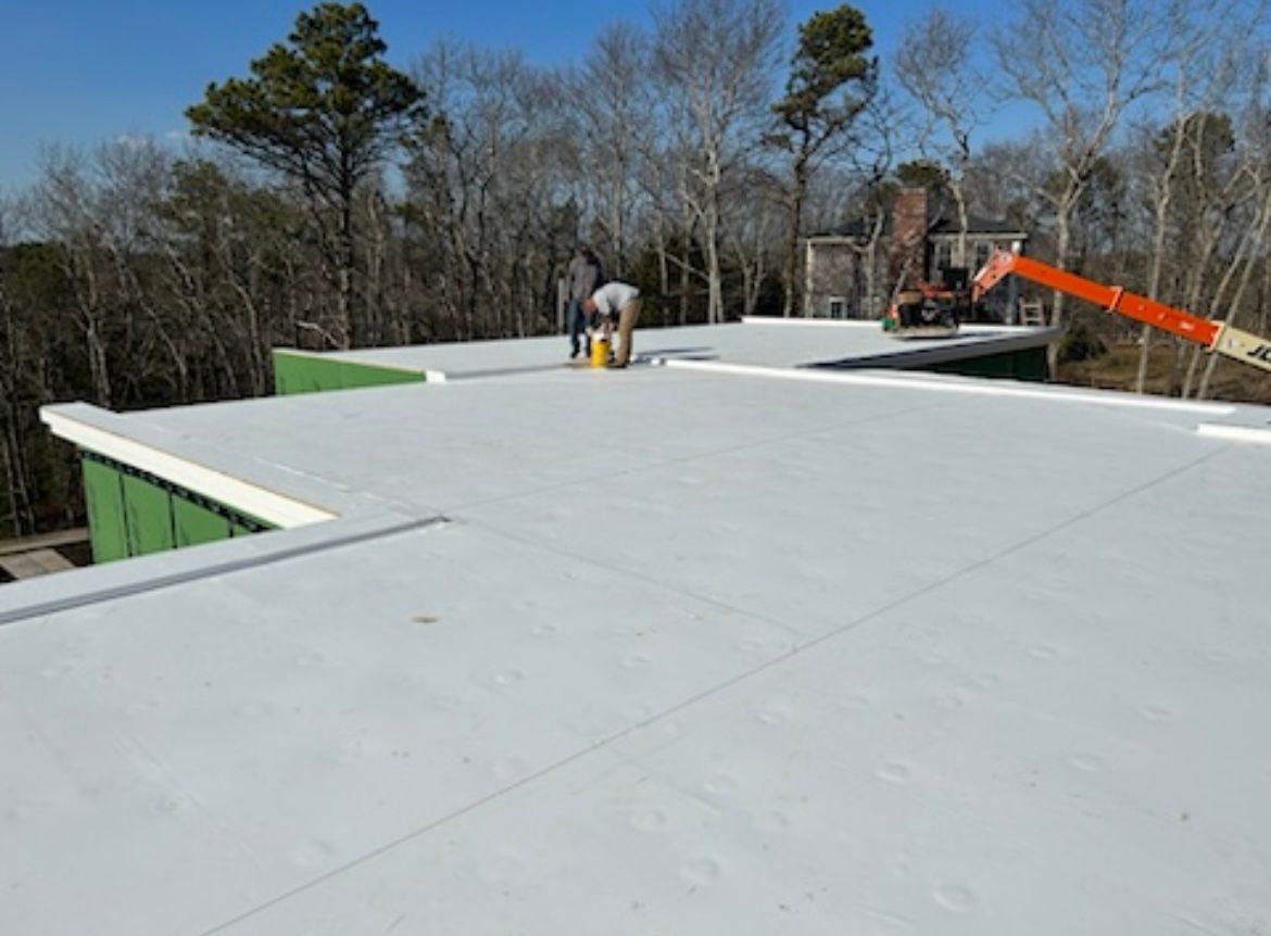 Workers on a flat white rooftop during construction, with a lift and bare trees in the background