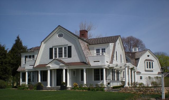 Large white farmhouse with a front porch, black shutters, and a green lawn in daylight