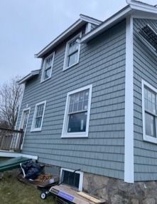 Gray two-story house with white trim and a basement window, viewed from the side in cloudy weather.