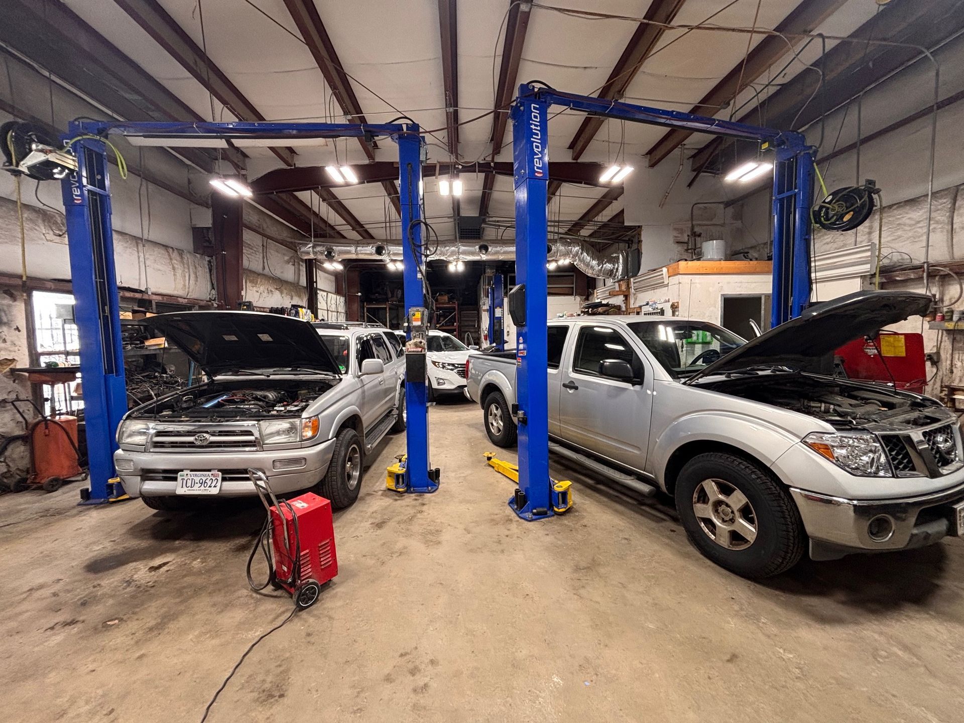 Two vehicles with open hoods in a garage, on lifts. Other vehicles visible.