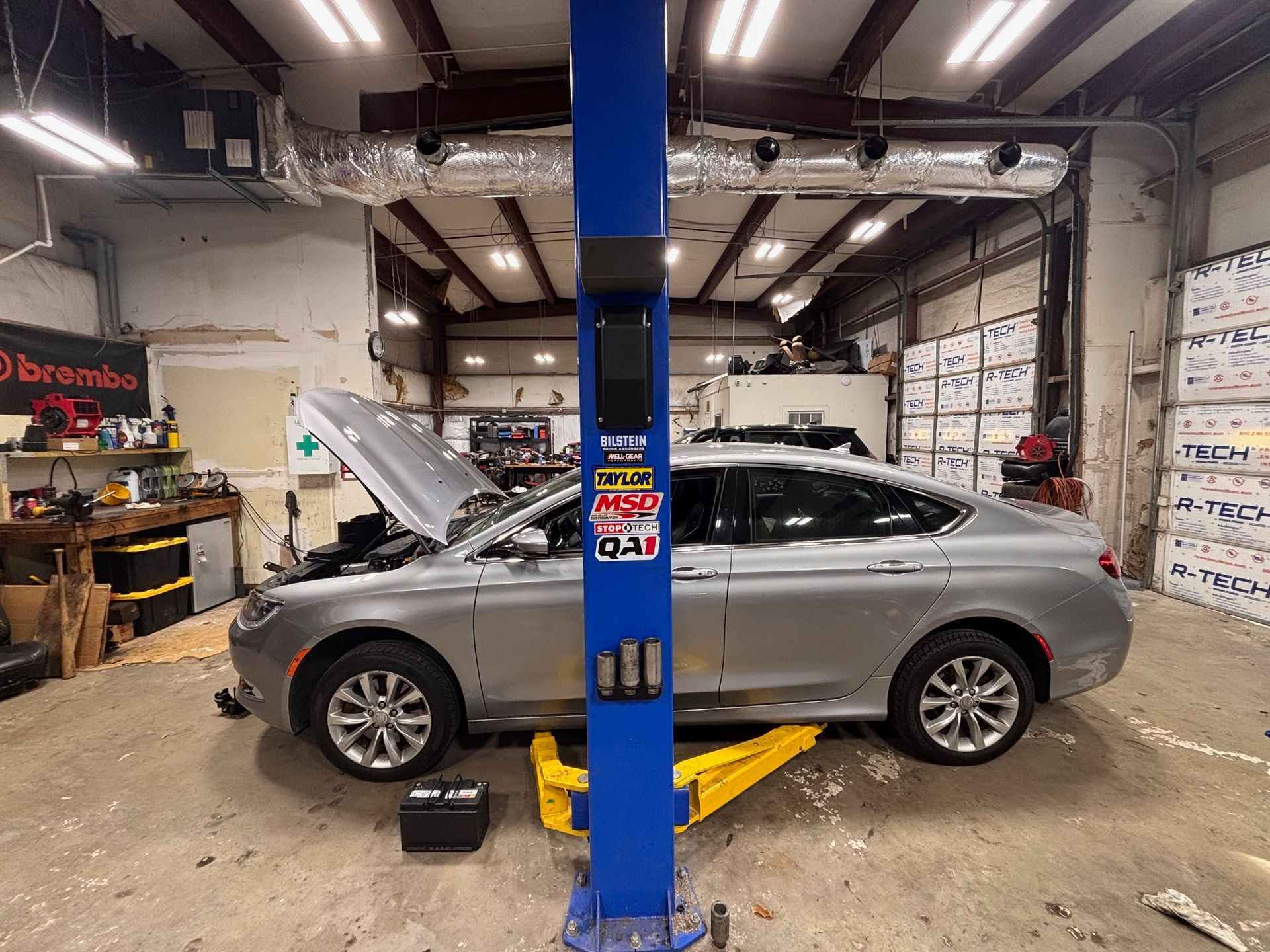Silver car on a lift in a garage with the hood open, blue lift support in the foreground.