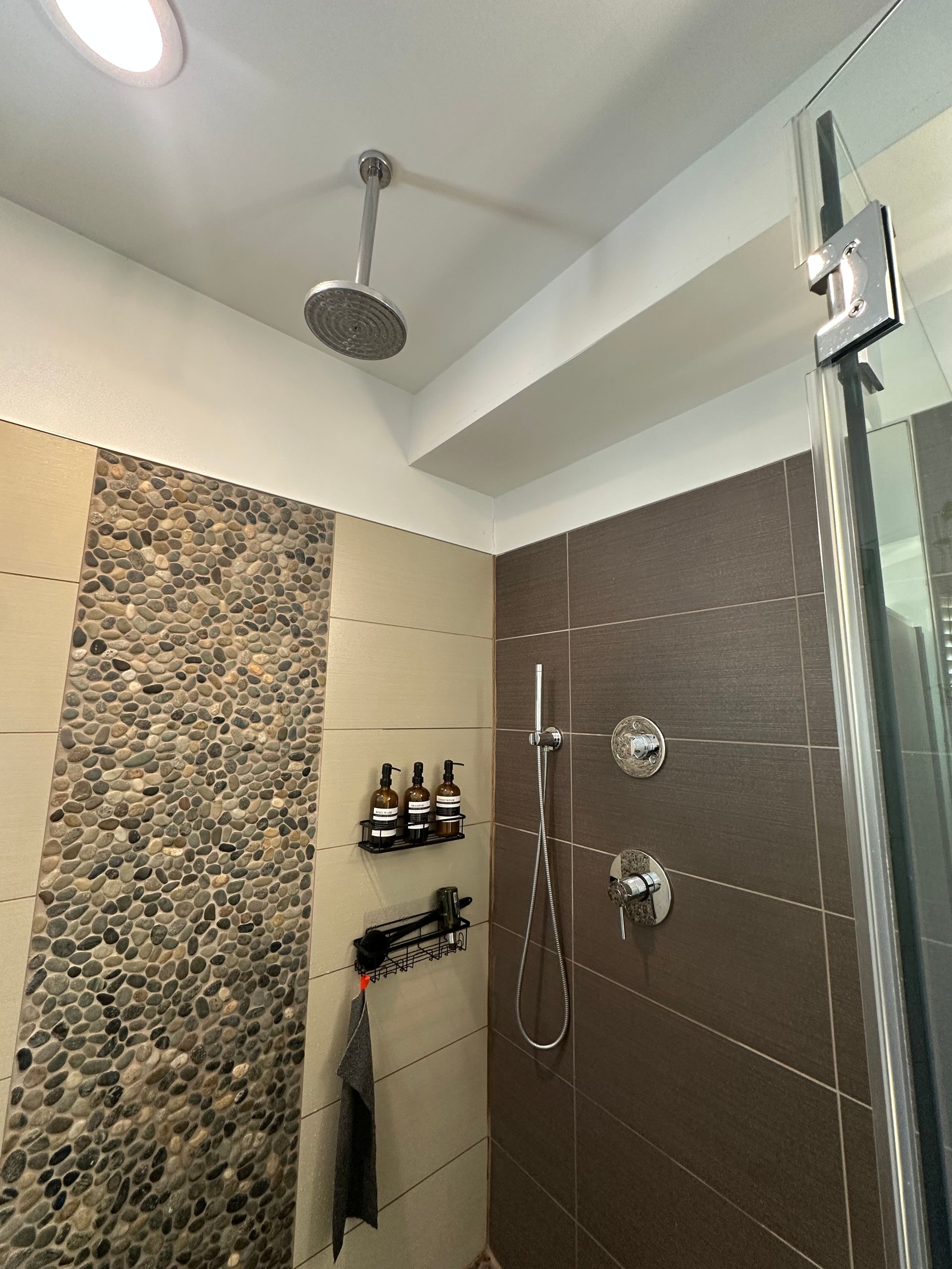 Modern shower with pebble accent wall, dark tile, glass door, and mounted shower fixtures.