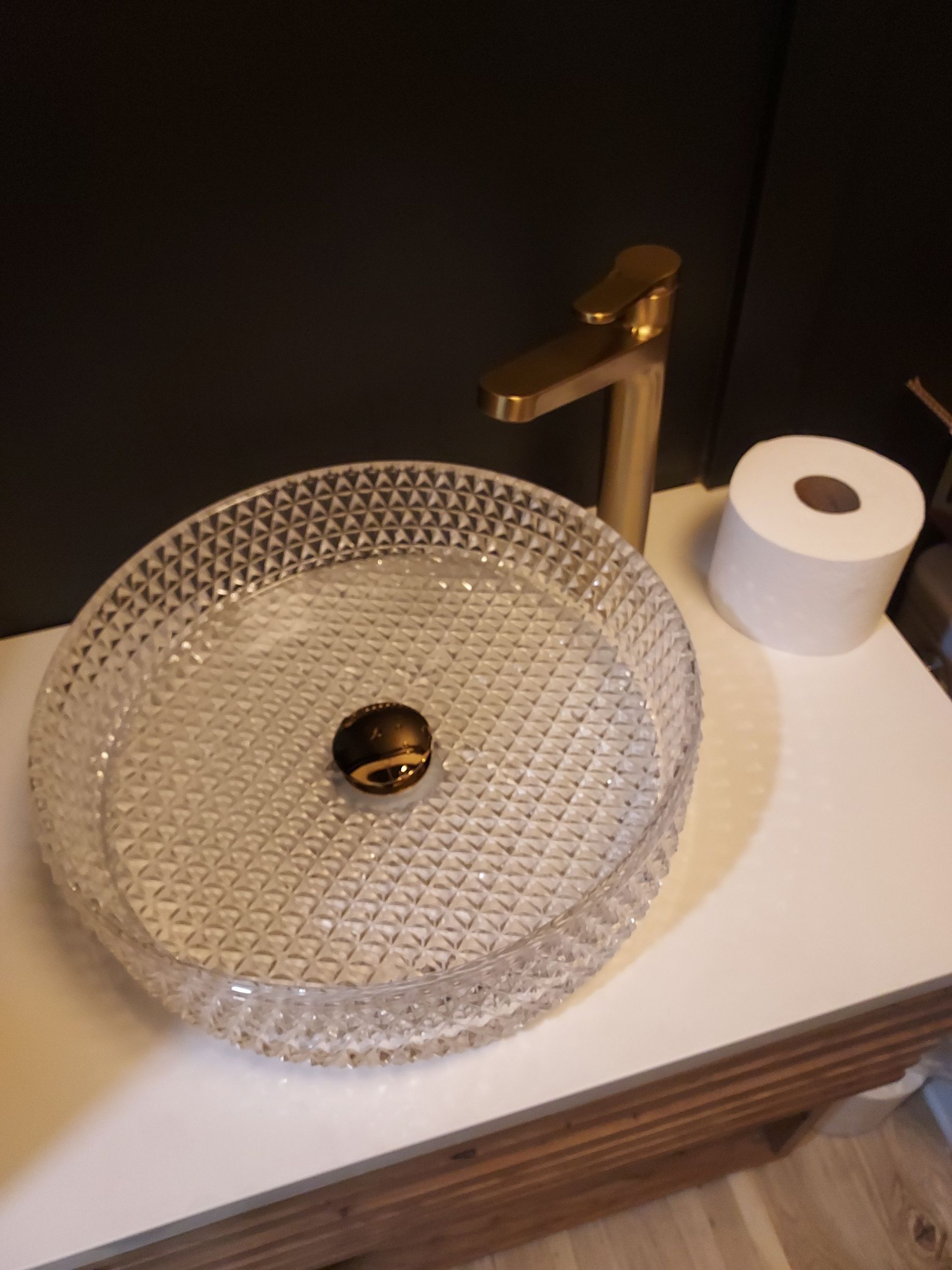 Glass vessel sink on a bathroom countertop with a gold faucet and toilet paper roll