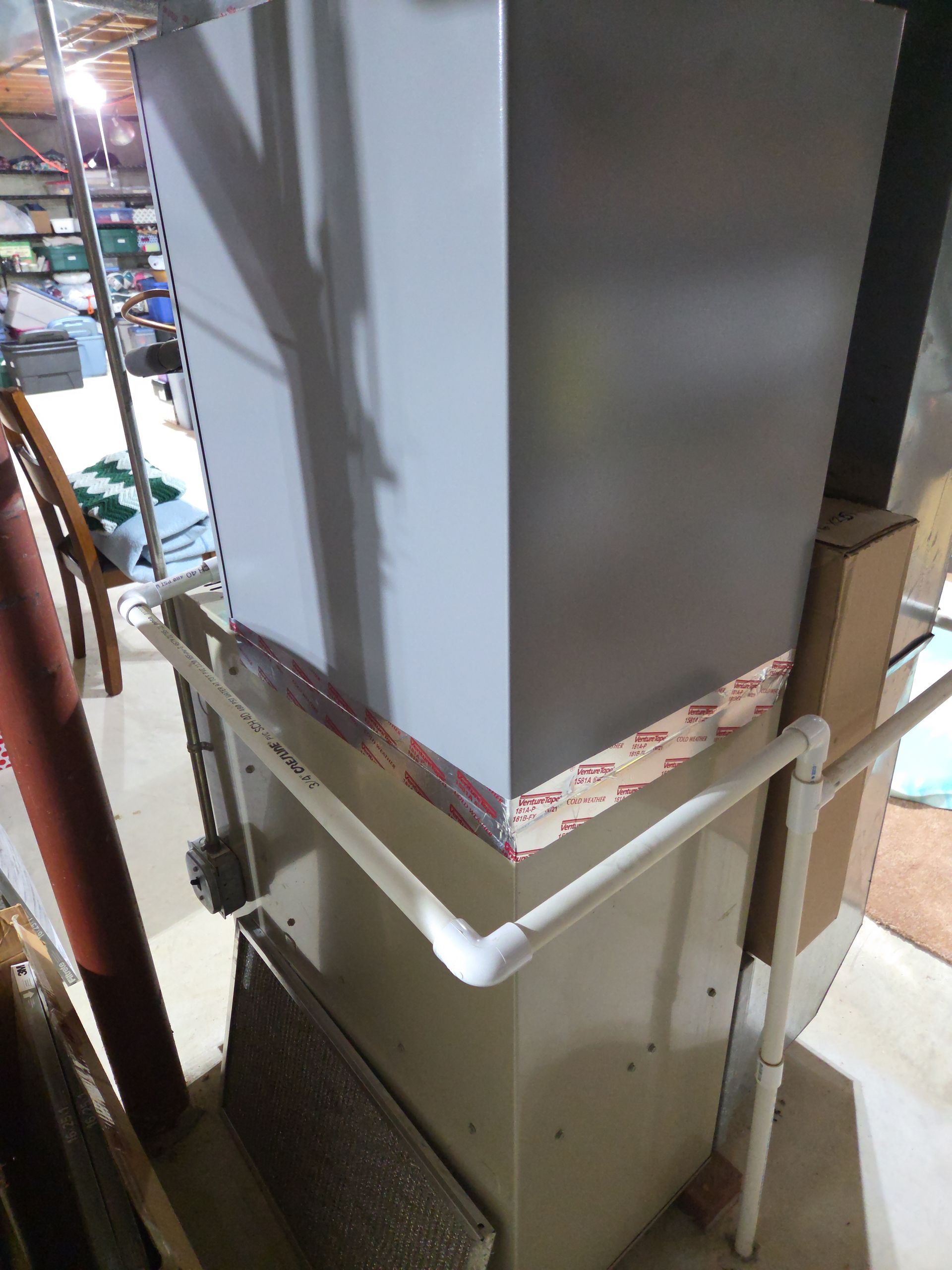 Large gray appliance on a beige cabinet in a storage area, with a taped seam and visible pipes below