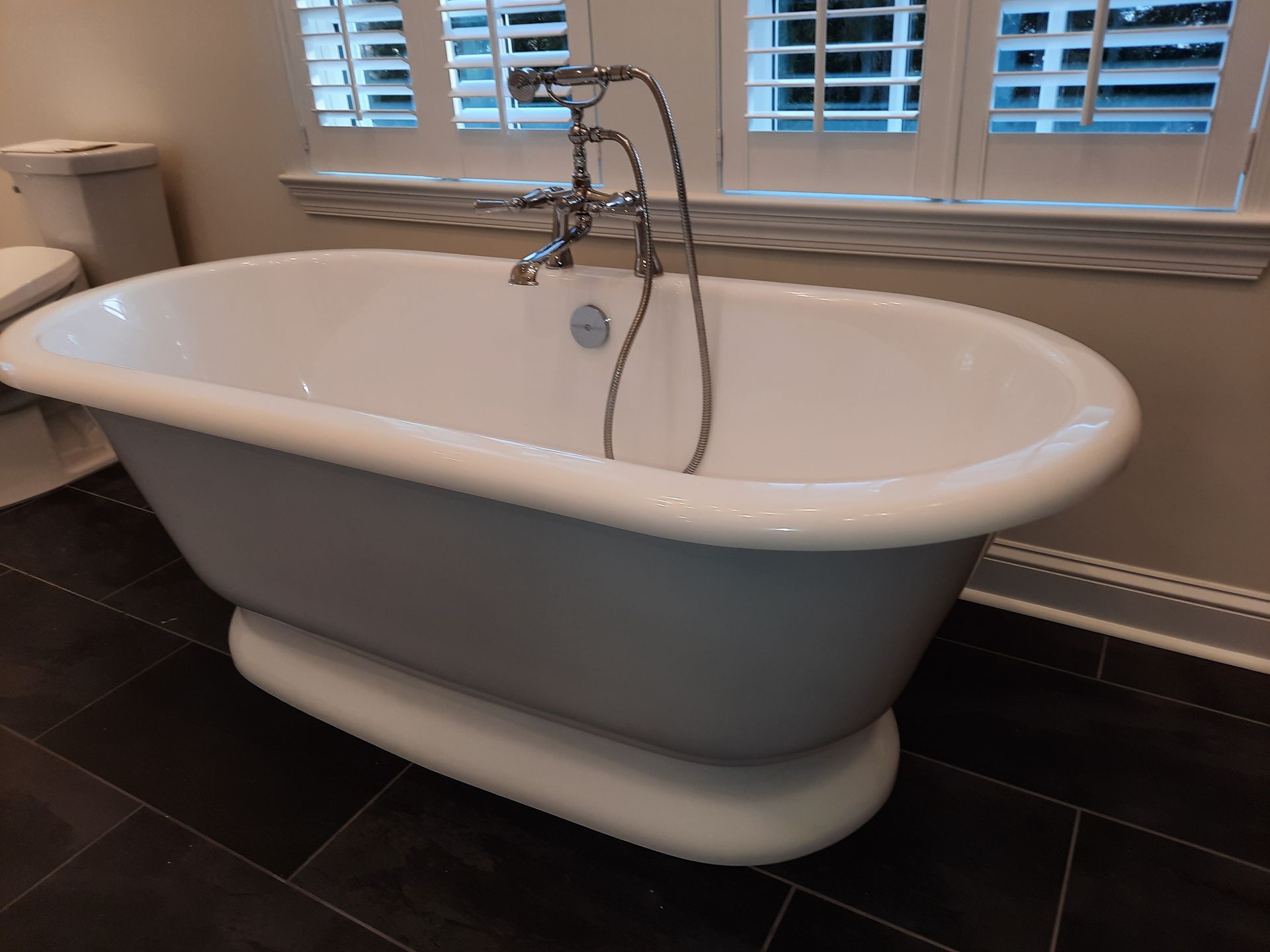 Freestanding white bathtub in a tiled bathroom with a chrome faucet and window shutters