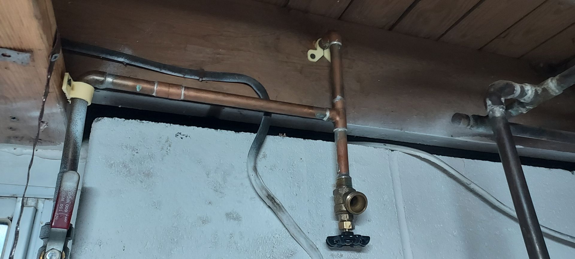 Exposed copper pipes and wall-mounted plumbing beneath a wooden ceiling in an unfinished room