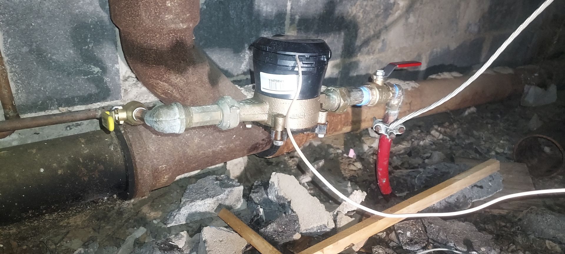 Plumbing under a sink with leaking pipes, a water meter, and red shutoff valve in a cluttered basement-like area