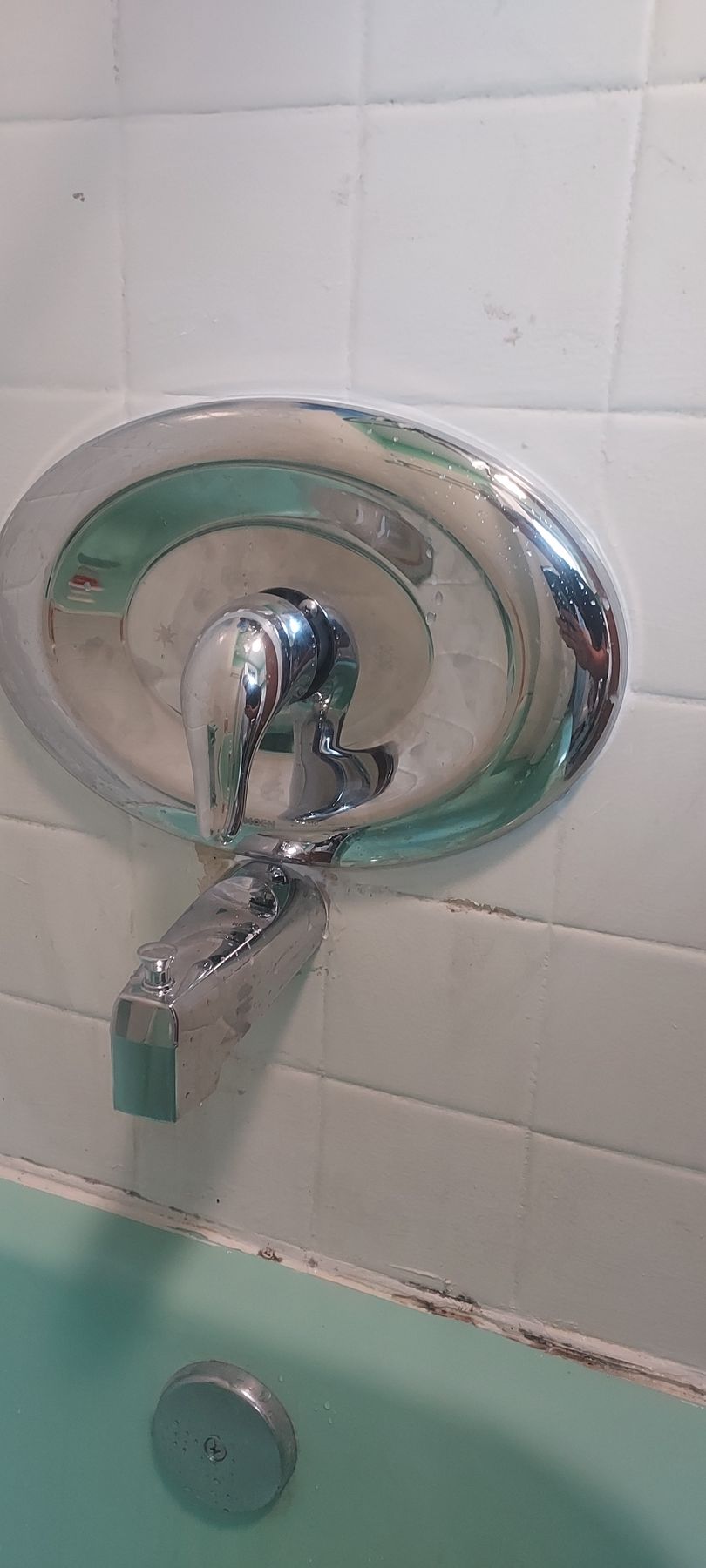 Chrome shower valve and faucet on white tile wall above a bathtub with mint-green water.