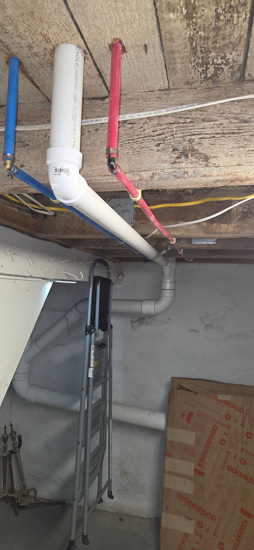 Exposed blue, white, and red plumbing pipes running along a basement ceiling near a utility area
