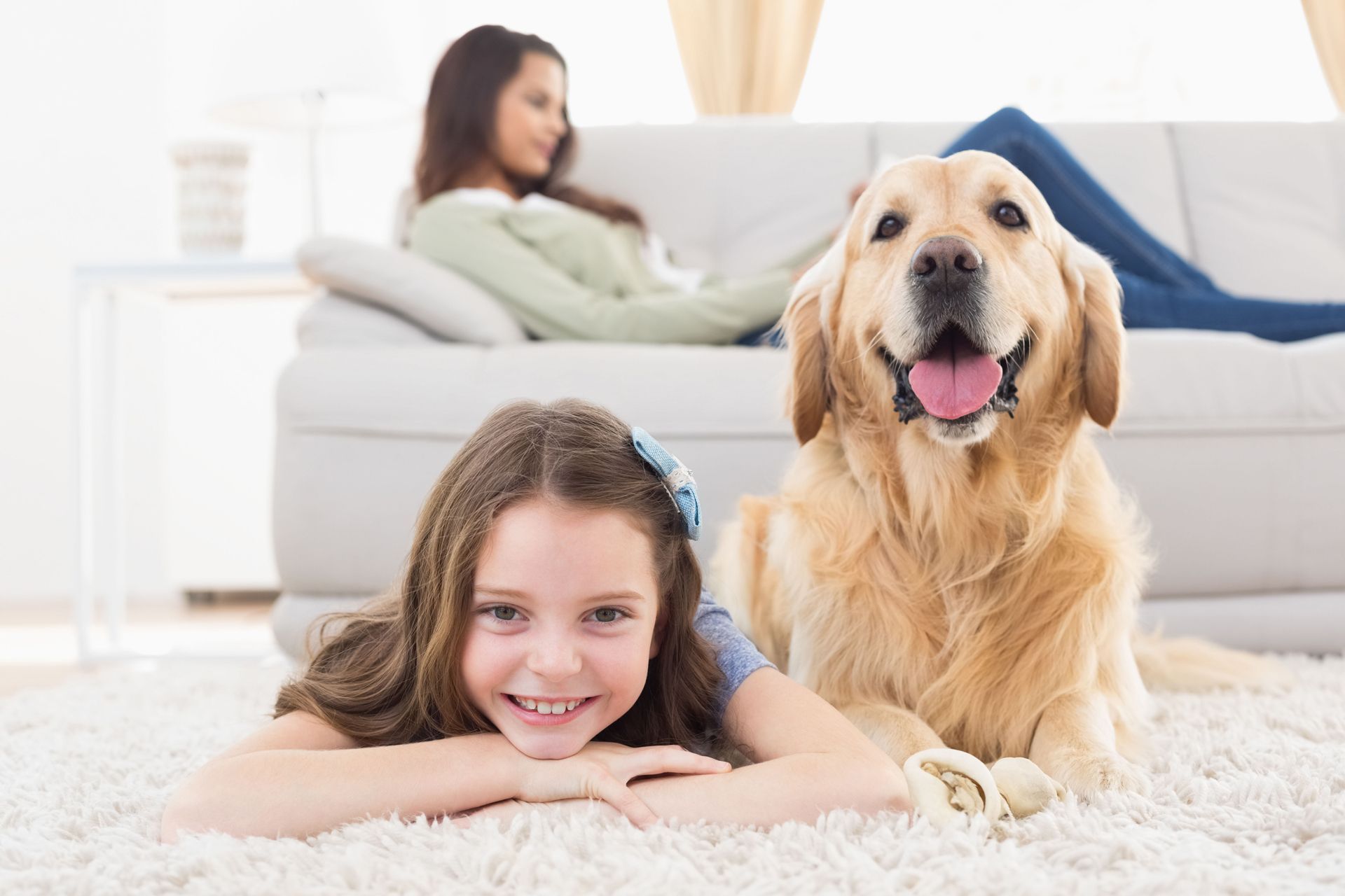 Girl lying on a fluffy rug beside a golden retriever, with a woman relaxing on a couch in the background.