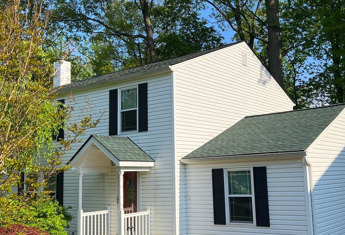 White two-story house with black shutters, green roof, and chimney, surrounded by trees.