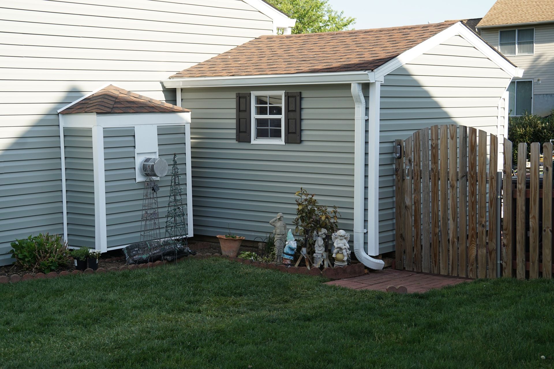 Green shed with brown roof, small window, and white trim next to a small white gazebo-like structure; wooden fence on right.